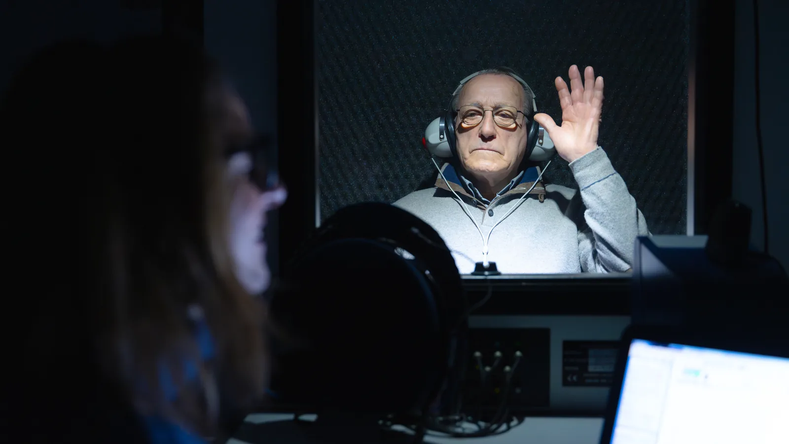 Homem com fones dentro de uma cabine audiométrica levantando a mão ao ouvir o som, durante a audiometria; a fonoaudióloga acompanha o exame.