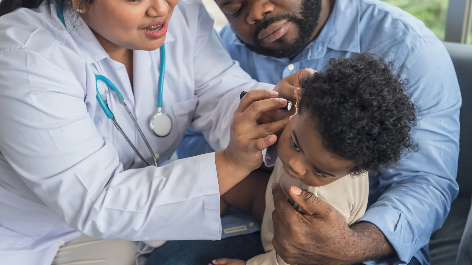 Médica examinando o ouvido de um bebê no colo do pai, em avaliação auditiva infantil.