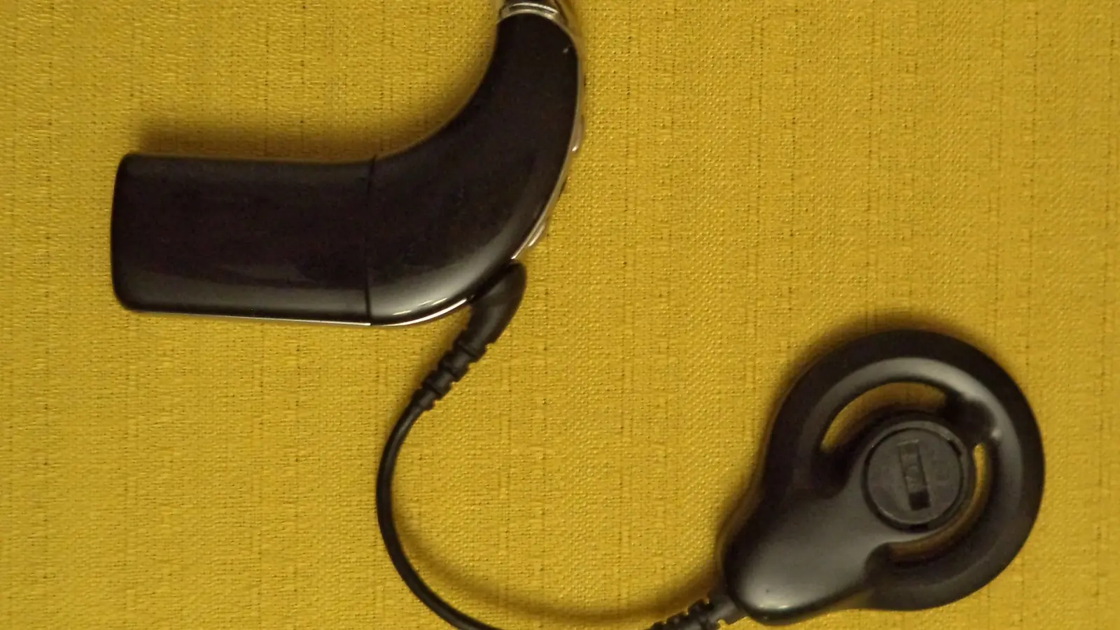 Unidade externa de um implante coclear vista de perto: o processador retroauricular ligado por um cabo à antena redonda que se fixa por ímã.