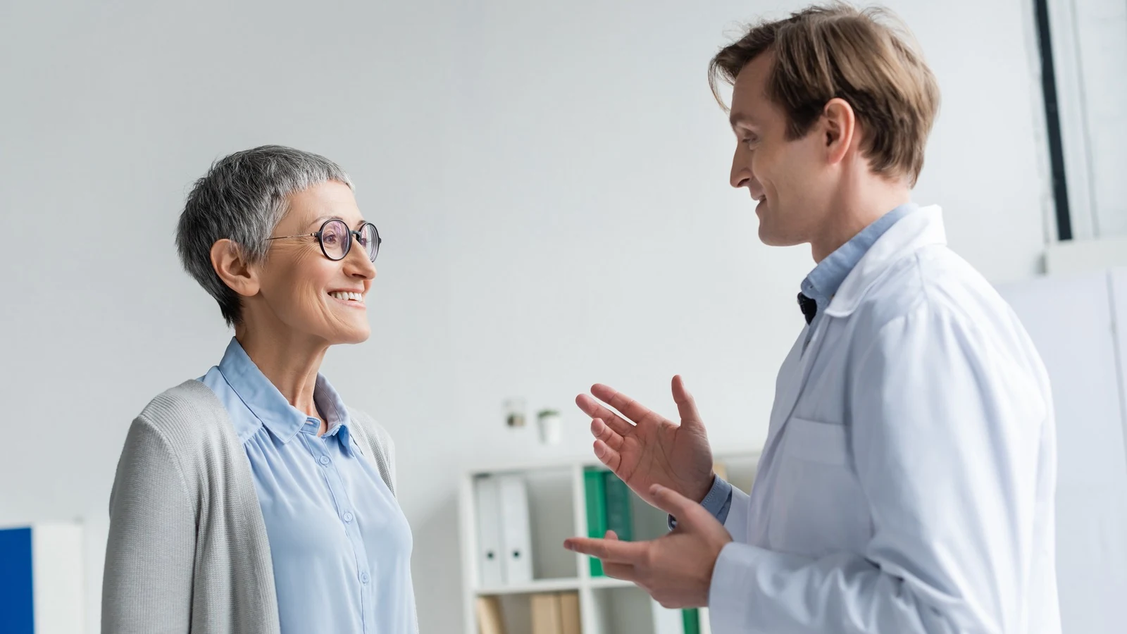 Médico de jaleco branco conversando com uma paciente de meia-idade no consultório, ela sorrindo enquanto ele explica gesticulando com as mãos.