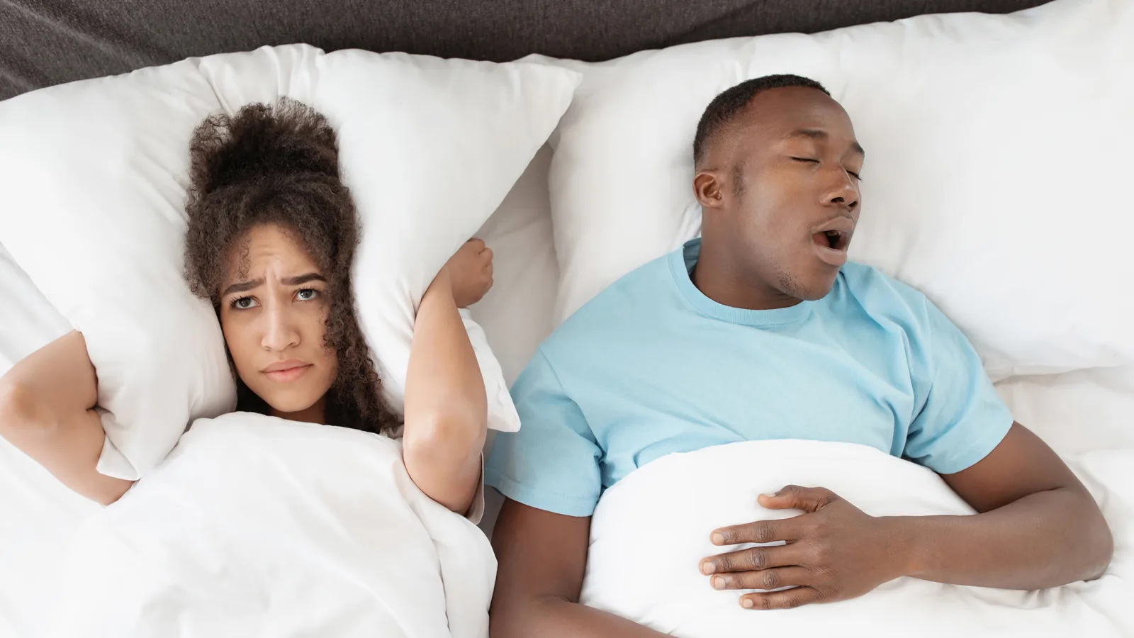 Casal na cama: o homem dorme de boca aberta roncando e a mulher, acordada, tapa os ouvidos com o travesseiro.